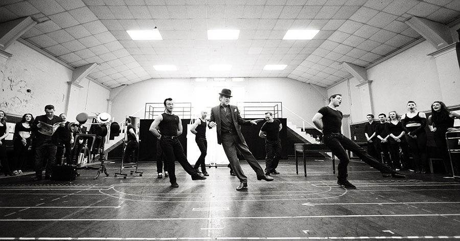 A wide photograph of the cast rehearsing for Dirty Rotten Scoundrels